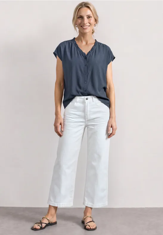 Vrouw in een blauw topje, witte wijde broek en zwarte sandalen tegen een neutrale achtergrond.