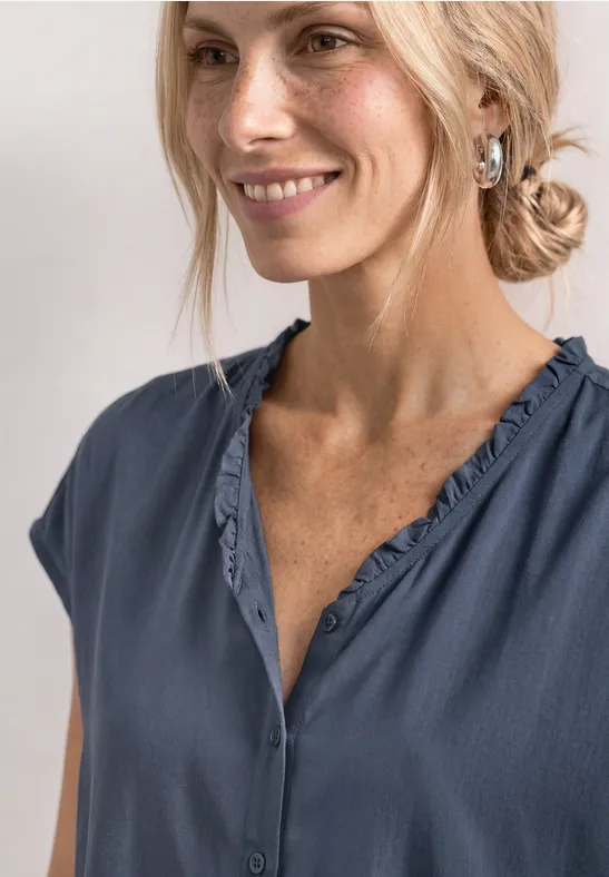 Blonde vrouw in een los blauw shirt met V-hals, glimlacht zachtjes opzij naar de camera.