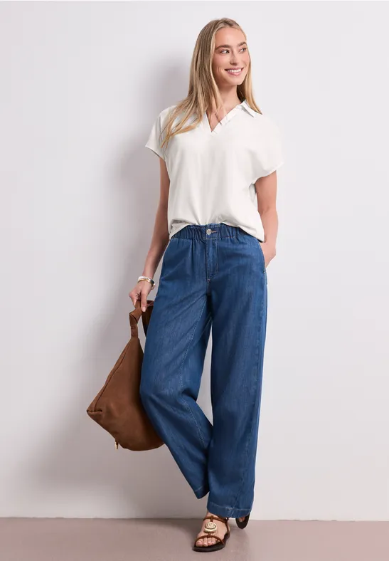 Eine Frau steht vor heller Wand, trägt weiße Bluse, weite Jeans, Sandalen und hält eine braune Tasche.
