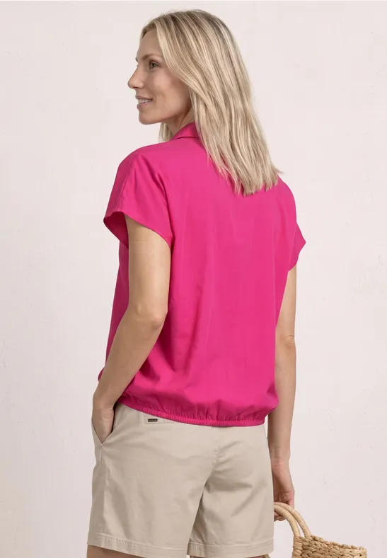 Frau von hinten in pinkem T-Shirt und beigen Shorts vor hellem Hintergrund.
