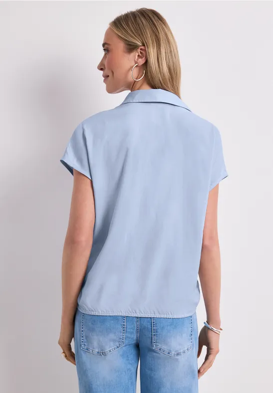 Frau von hinten in hellblauer Kurzarmbluse und Jeans vor neutralem Hintergrund.