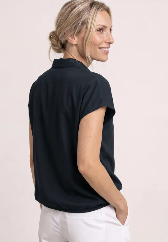 Vue arrière d'une femme portant une chemise à manches courtes noire et un pantalon clair, mains dans les poches.