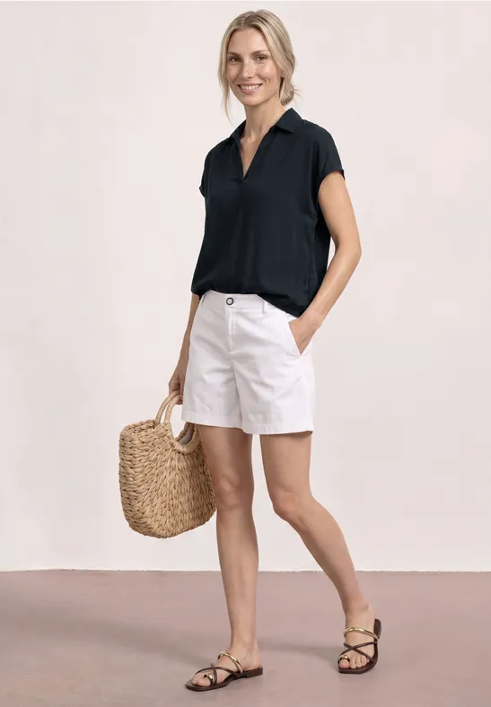 Femme en chemisier noir, short blanc, sac en paille et sandales fines noires sur fond clair.