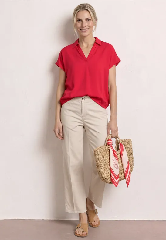 Frau in roter Bluse, weißer Hose und Basttasche mit Schal vor hellem Hintergrund.