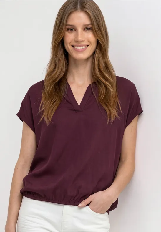 Femme en chemisier bordeaux à manches courtes et pantalon blanc, debout main dans la poche.