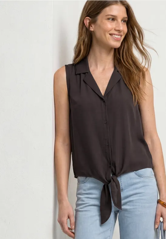 Eine junge Frau trägt eine ärmellose, knöpfbare Bluse mit Knoten vorne und eine helle Jeans.