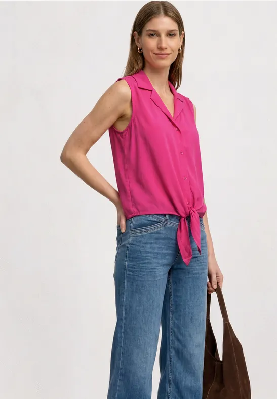 Frau in ärmelloser, pinker Bluse mit Knoten vorne, blauen Jeans und brauner Tasche, seitlich stehend.