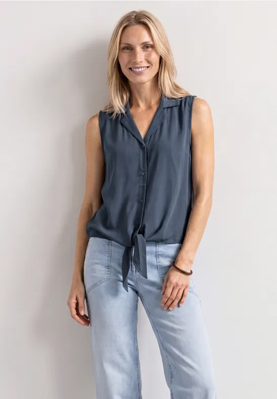 Eine blonde Frau lehnt vor weißer Wand, trägt ärmellose dunkle Bluse mit Knoten und helle Jeans.