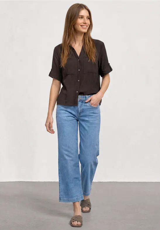 Frau in schwarzem kurzärmeligem Hemd, weiten blauen Jeans und Sandalen vor neutralem Hintergrund.