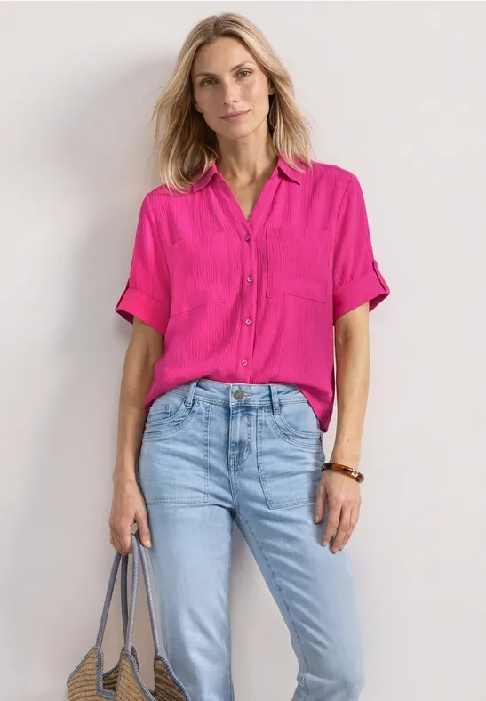 Vrouw met roze blouse en lichte jeans, grijze tas in hand, tegen lichte achtergrond.