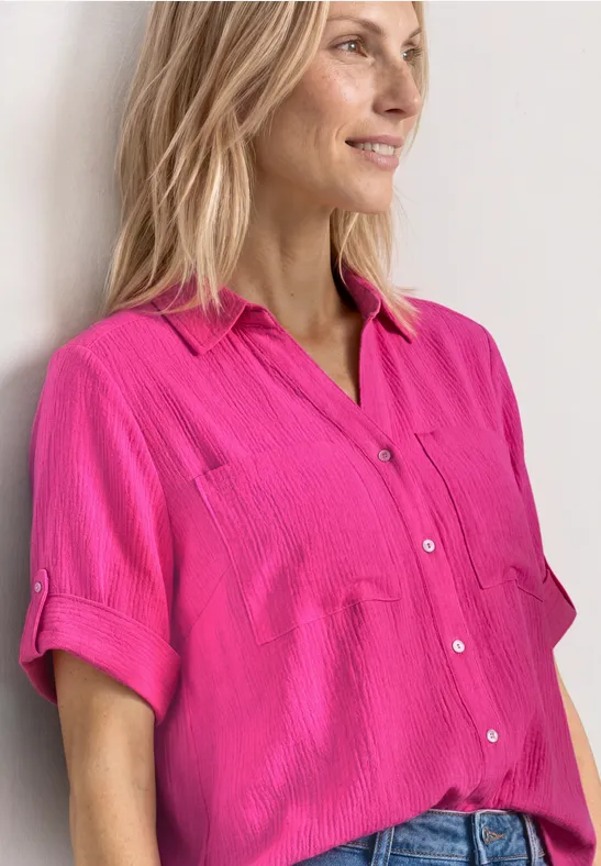 Blond persoon in roze overhemd met korte mouwen leunt tegen een witte muur.