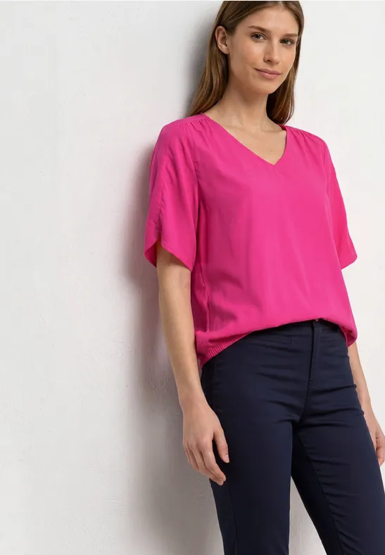 Persoon staat tegen een witte muur, draagt een roze shirt en een donkere broek.