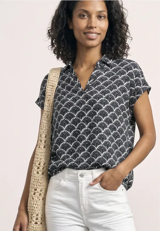 Eine Person trägt eine schwarze Bluse mit kurzem Arm und Muschel-Print, weiße Jeans und eine gehäkelte Strohtasche.