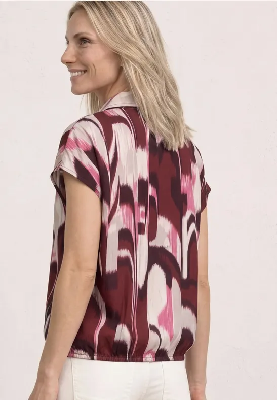 Eine Frau von hinten mit blondem Haar trägt eine kurzärmelige Bluse mit pink-weiß-bordeauxfarbenem Muster.