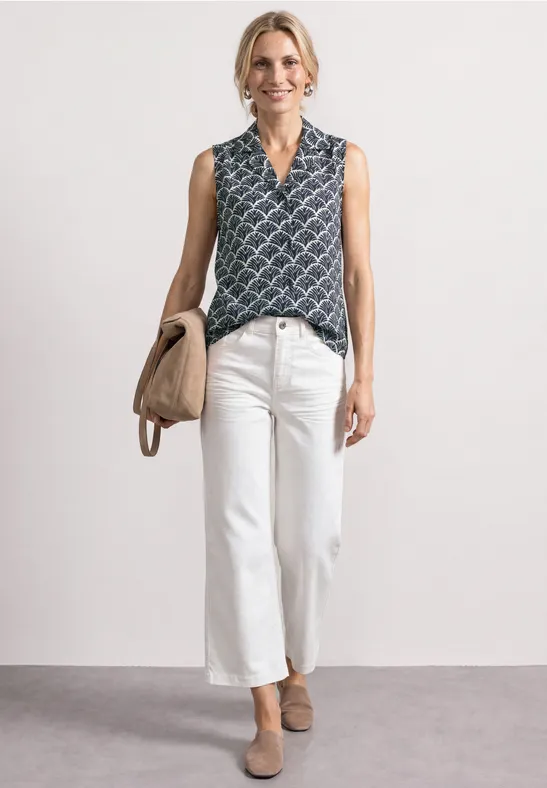Frau in gemusterter ärmelloser Bluse, weißen Culotte-Jeans, beigen Pumps und passender Tasche.