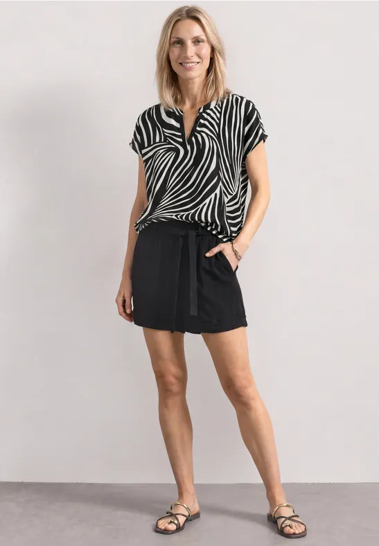 Vrouw in zebrapatroon blouse, korte zwarte rok en sandalen tegen een neutrale achtergrond.