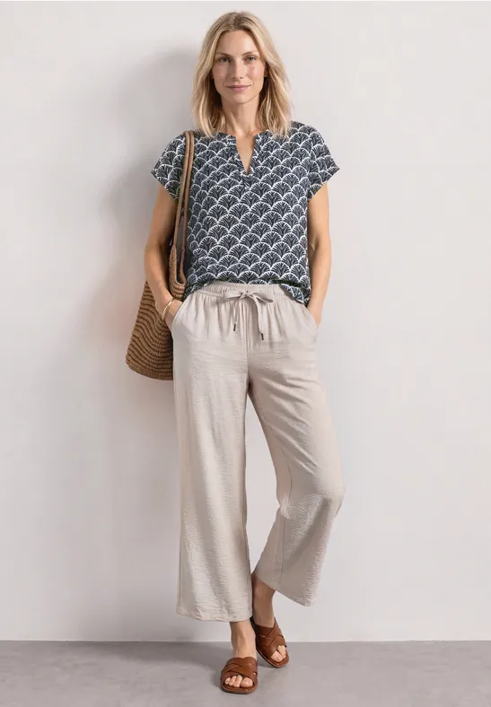 Frau trägt blaue kurzärmlige Bluse mit weißem Muster, beige weite Hose, braune Sandalen und geflochtene Tasche.
