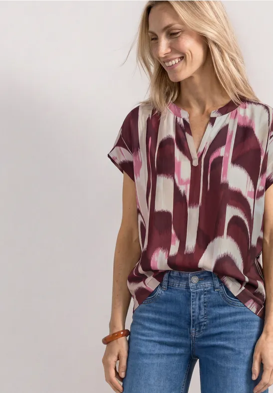 Vrouw draagt losvallende korte mouw blouse met abstract rood-wit dessin en jeans.
