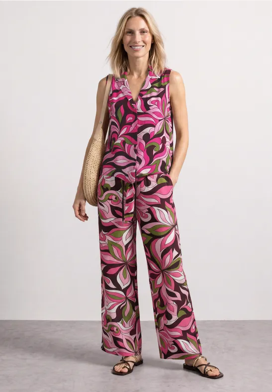 Vrouw draagt mouwloze, roze bloemenprint top met wijde pantalon, gecombineerd met sandalen en een rieten tas.