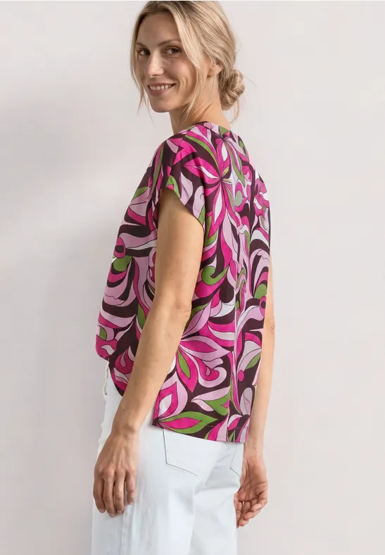 Vrouw in kleurrijke, korte mouwen top met abstracte swirlprint in roze, groen en zwart en witte broek.