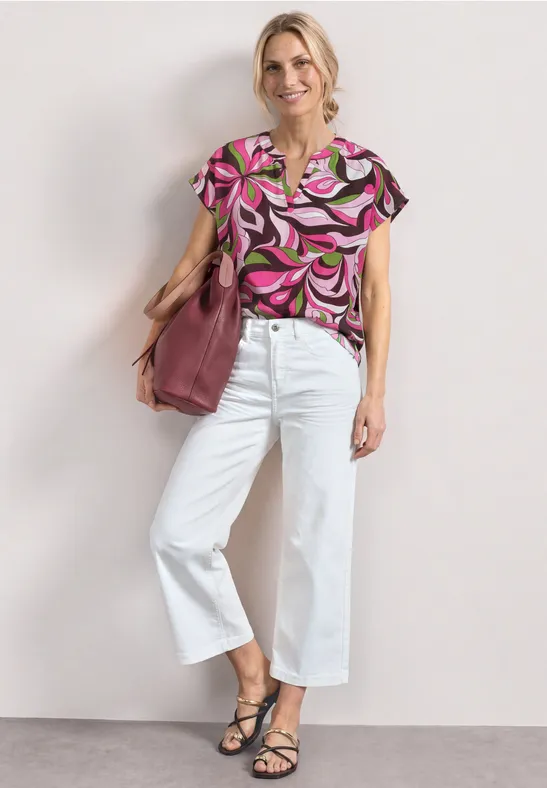 Vrouw met knot in roze dessinblouse, witte wijdere broek, zwarte sandalen en een grote roze tas.