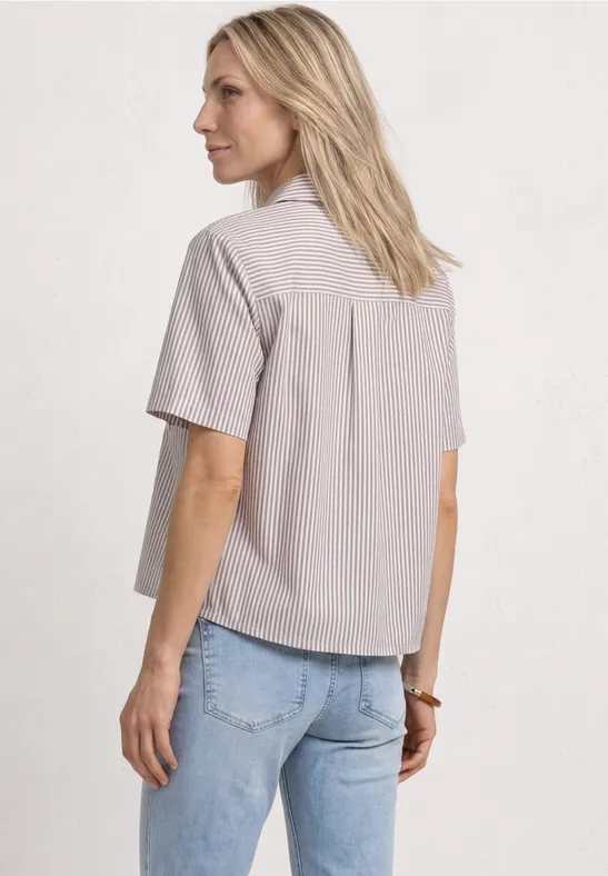 Frau in hell gestreiftem Kurzarmhemd und blauer Jeans, von hinten vor neutralem Hintergrund.