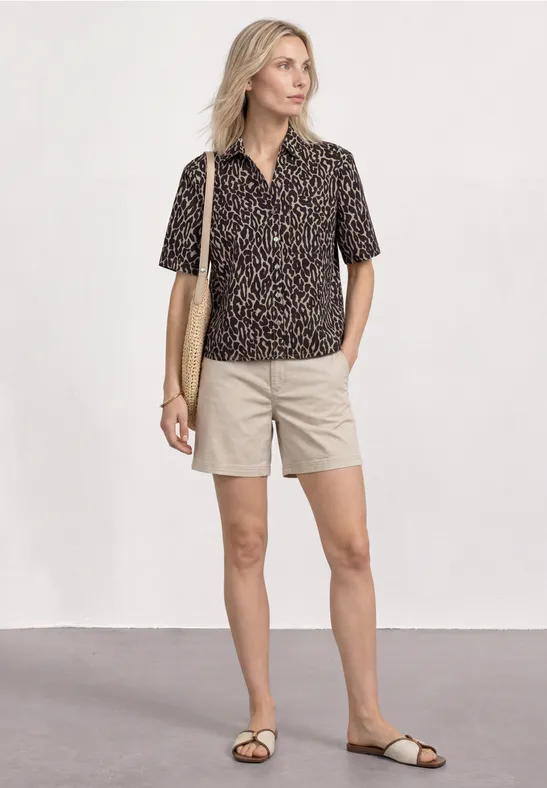 Vrouw met luipaardprintblouse, beige short en sandalen, schoudertas, staand voor een neutrale achtergrond.