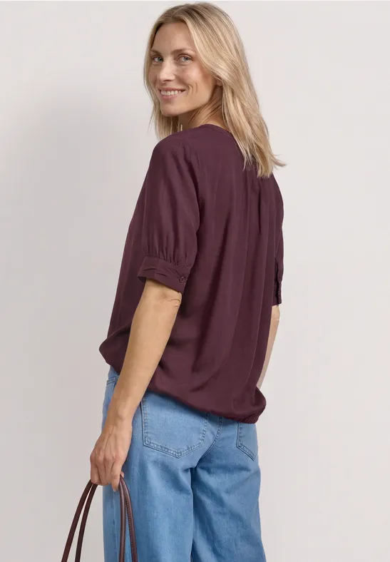 Een blond persoon van achter gezien met een paars shirt, blauwe jeans en een tas in de hand.