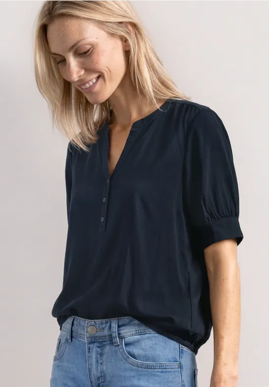 Vrouw lacht in een donkerblauw hemd en jeans, staand voor een lichtgrijze achtergrond.