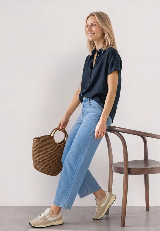 Vrouw in marineblauw overhemd en lichte jeans, gevlochten tas en gouden sneakers, staand naast een stoel.