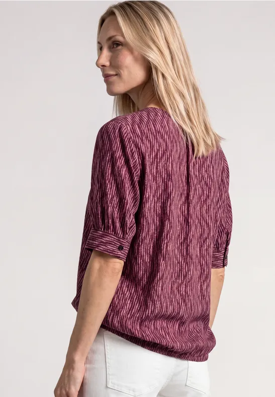 Achteraanzicht van een vrouw met blond haar in een paarse kabeltrui en witte broek voor een lichte achtergrond.