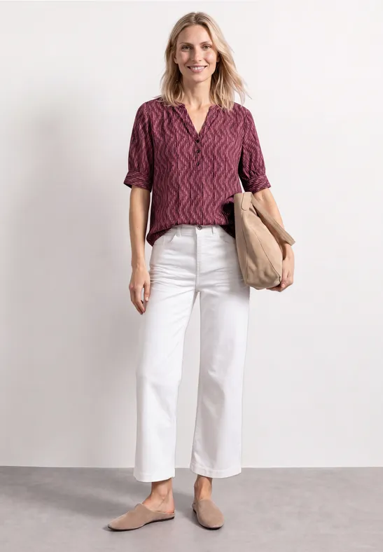 Vrouw in een bordeauxrood patroon-top, witte wijde broek, beige slippers en een taupekleurige tas.