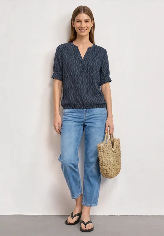 Vrouw in T-shirt met patroon, blauwe jeans, slippers en rieten tas voor neutrale achtergrond.