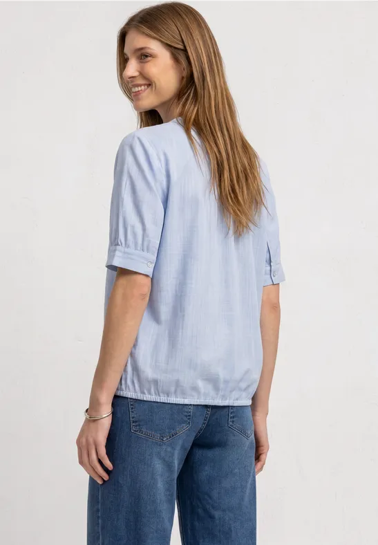 Eine Frau in hellblauer Bluse und Jeans, seitlich gewandt und lächelnd.