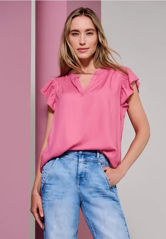 Frau in rosa Bluse und Jeans posiert vor zweifarbigem Hintergrund.