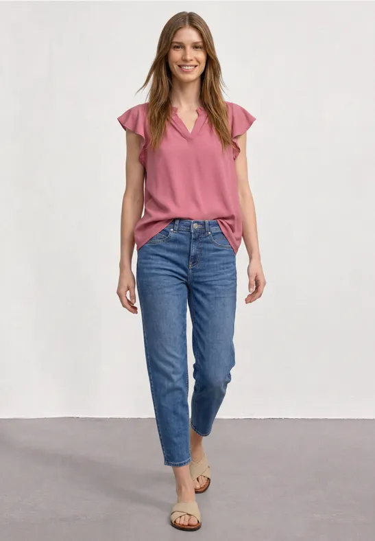 Junge Frau in Jeans und rosa Top, steht lächelnd in Studio.