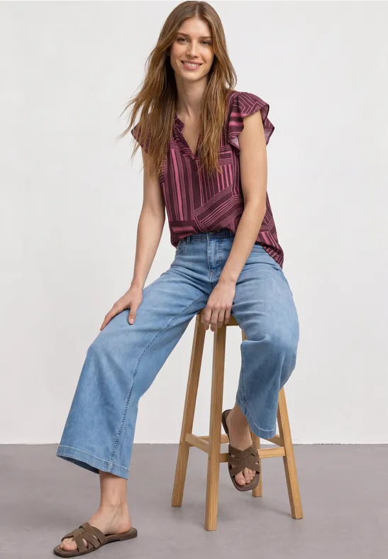 Een vrouw zit op een kruk, draagt een gestreept topje, wijde jeans en sandalen.