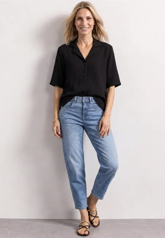 Frau in schwarzer Bluse, Jeans und Sandalen vor neutralem Hintergrund.