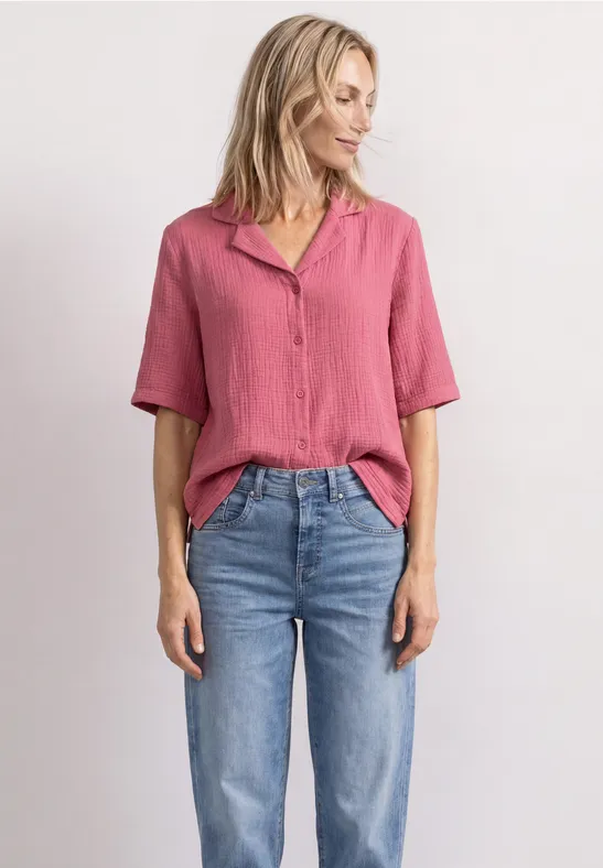Frau in rosa Bluse und blauen Jeans steht lässig vor weißem Hintergrund.