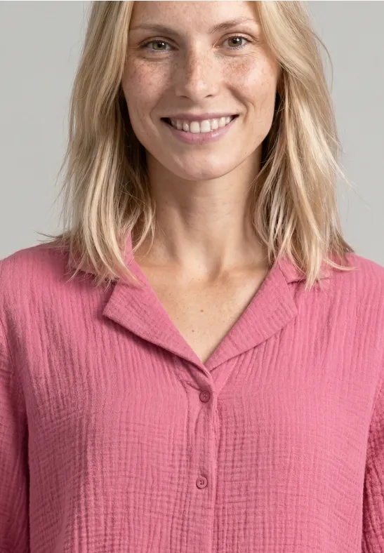 Eine lächelnde Frau mit blondem Haar trägt eine rosa, strukturierte Bluse.