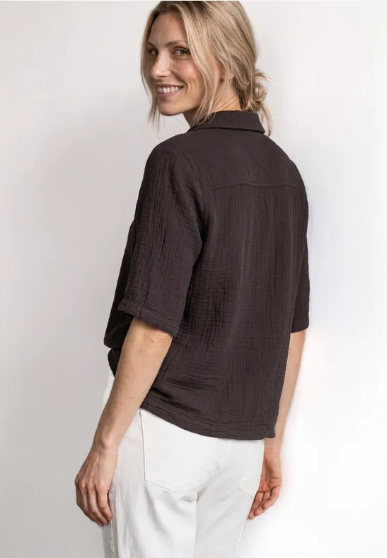 Blonde vrouw in losse bruine blouse en witte broek, kijkt lachend over haar schouder.