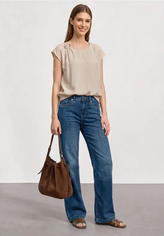 Frau mit beige Top, blauen Jeans, brauner Tasche und Sandalen vor weißem Hintergrund.