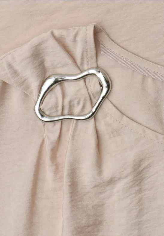 Nahaufnahme eines beigen Oberteils aus zartem Stoff mit unregelmäßigem silbernem Ring an der Schulterpartie.