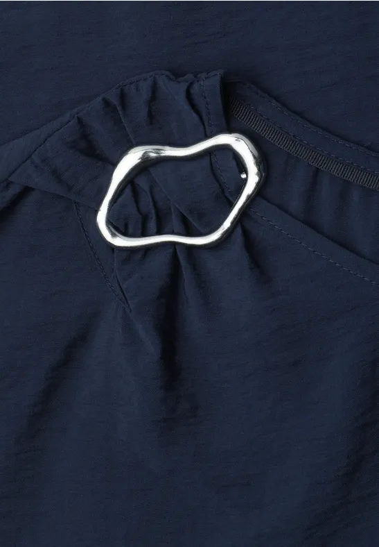 Dunkelblauer Stoff mit silbernem, organisch geformtem Ring als Zierdetail an einer Raffung.