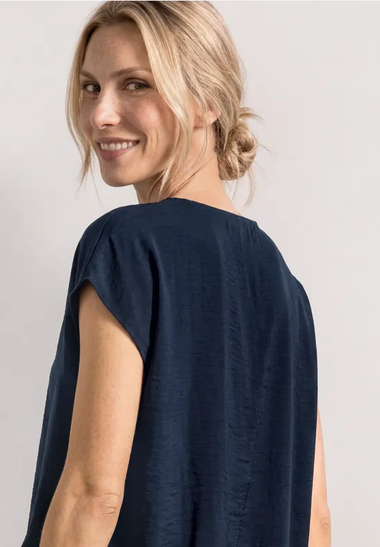 Rückenansicht einer Person mit blonden, tief gebundenen Haaren und dunkelblauem, kurzärmligem Oberteil.