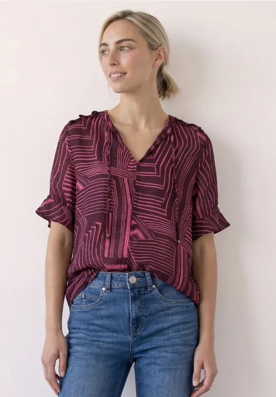 Vrouw in bordeaux-zwart gestreepte blouse met driekwartmouwen, los in jeans gestopt, voor een lichte muur.