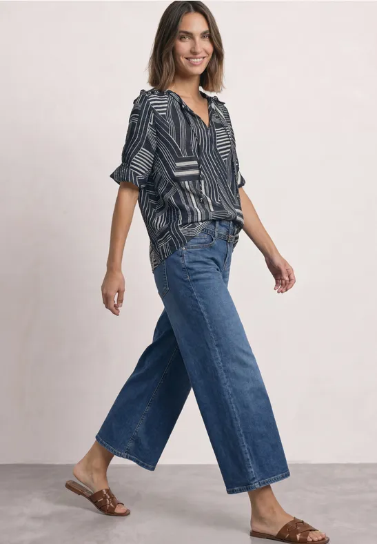 Vrouw in een gemêleerd shirt, wijde cropped jeans en platte sandalen tegen neutrale achtergrond.