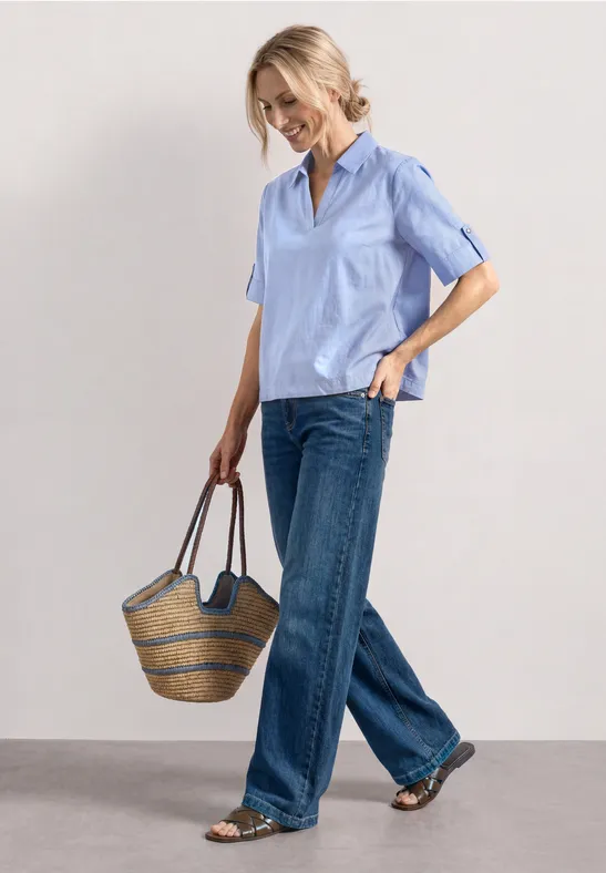 Frau in hellblauem Kurzarmhemd, weiten Jeans und Basttasche, entspannt im Gehen.