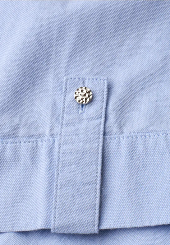 Detail eines hellblauen Hemdplackets mit einem verzierten Metall-Druckknopf.