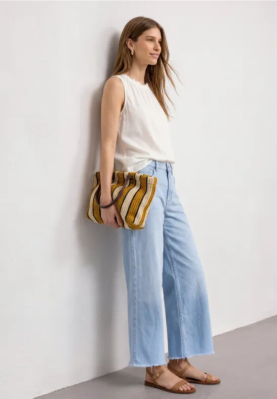 Frau in weißem Top, hellblauen weiten Jeans und braunen Sandalen lehnt an weißer Wand und hält gestreifte Clutch.